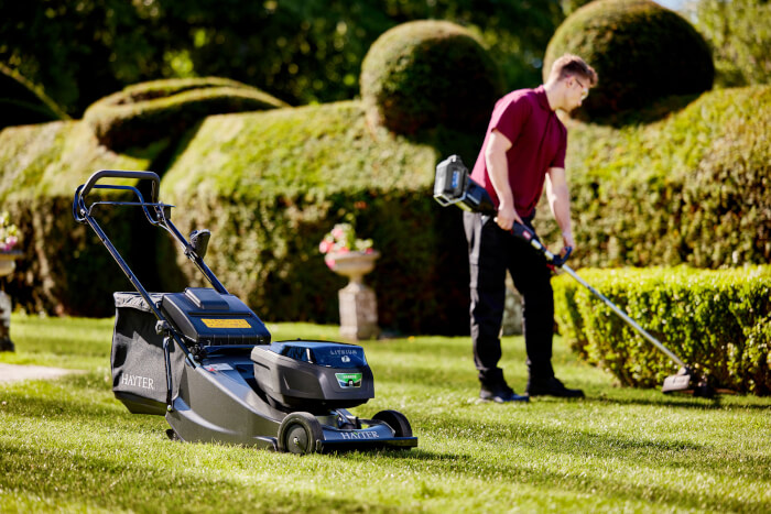 Hayter Harrier 48 Pro cordless lawnmower on a freshly cut lawn, with a professional landscaper using a battery-powered trimmer to shape hedges in the background